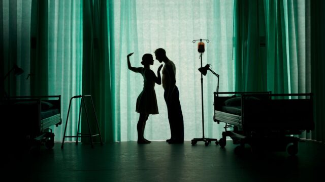 A hospital scene. Two dancers are standing close together face to face. They are behind a hospital curtain so you can only see their shadows.