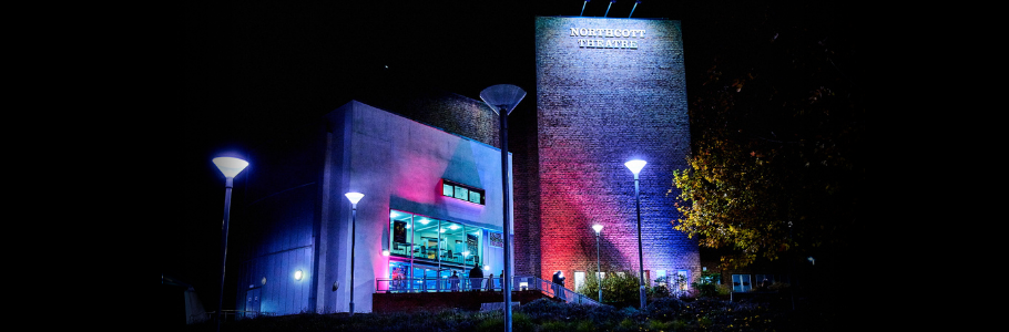 Image of Exeter Northcott Theatre taken at night. The sky is black and the building is lit with red and blue lights. On the right of the building, a sign reads 'Exeter Northcott Theatre'. Silhouettes of people appear in front of the building.