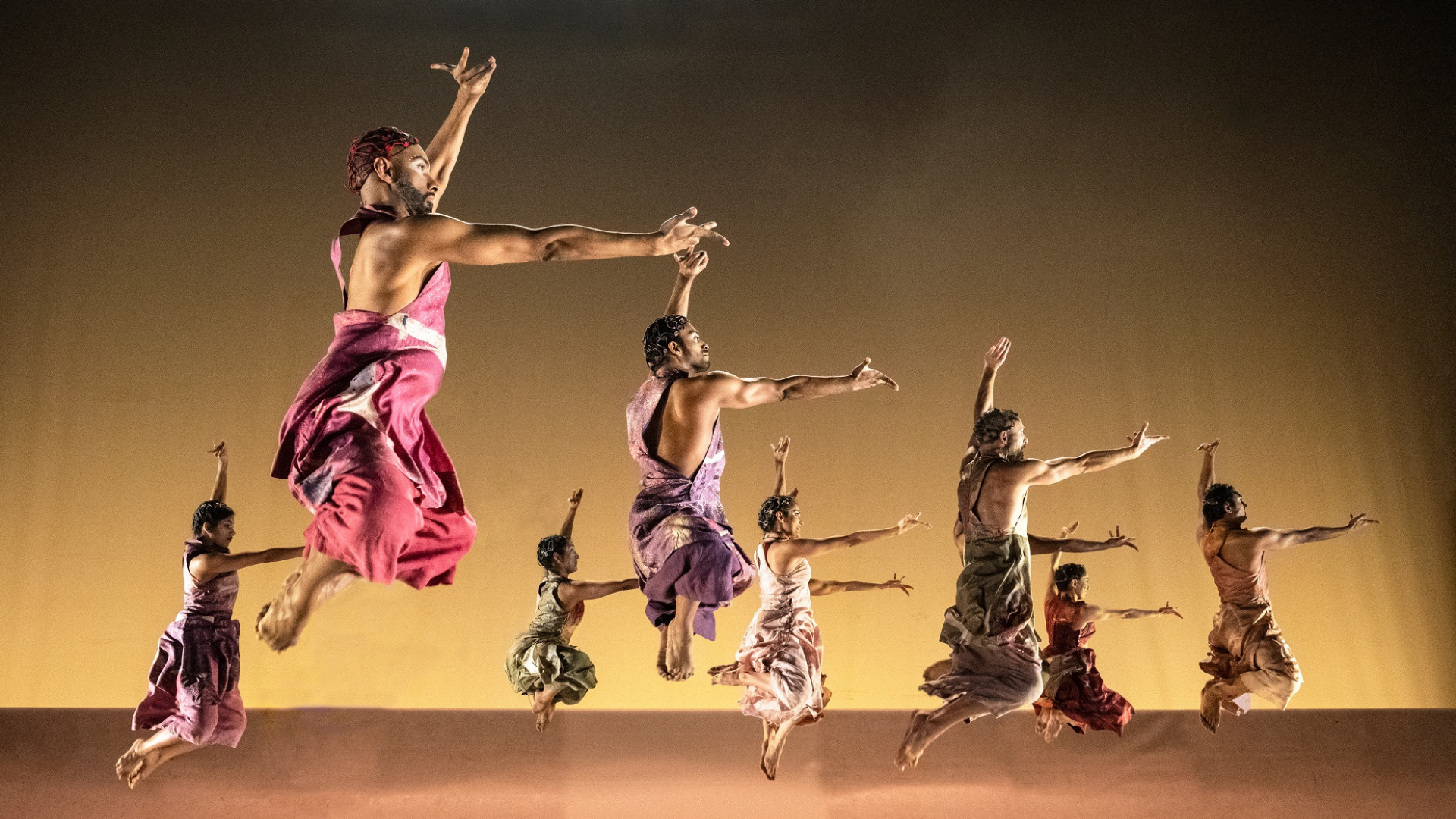 Production shot of The Rite of Spring. Two rows of dancers leap into the air with their arms outstretched and their knees bent.