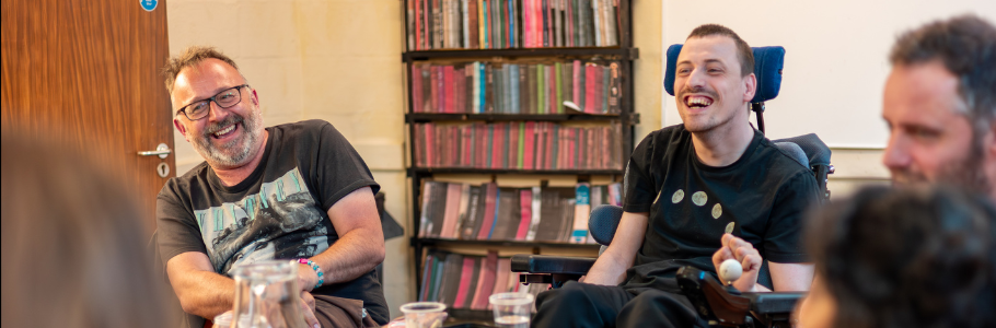 A person sits next to Hugh Malyon in a room in front of a bookcase. They both appear to be laughing.