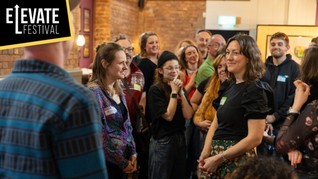 A group of Failspace participants stand and listen intently to another participant in the Barnfield Theatre's bar.