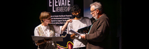 Image of 3 artists stood on a stage. On the left, the artist holds a book and points at another. The artists in the middle and right look down at their books. Behind them is a roller banner that reads 'Elevate Membership'.