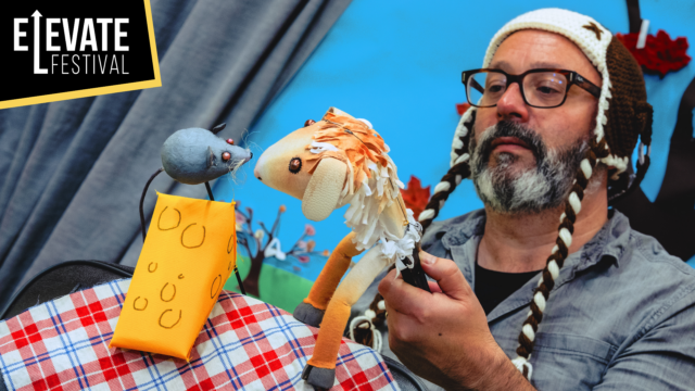 The Thing About Goats artwork. Shows a man with a knitted hat and beard, Holding a horse puppet. He is bringing the puppet close to a puppet of a mouse sitting on a triangle of cheese.