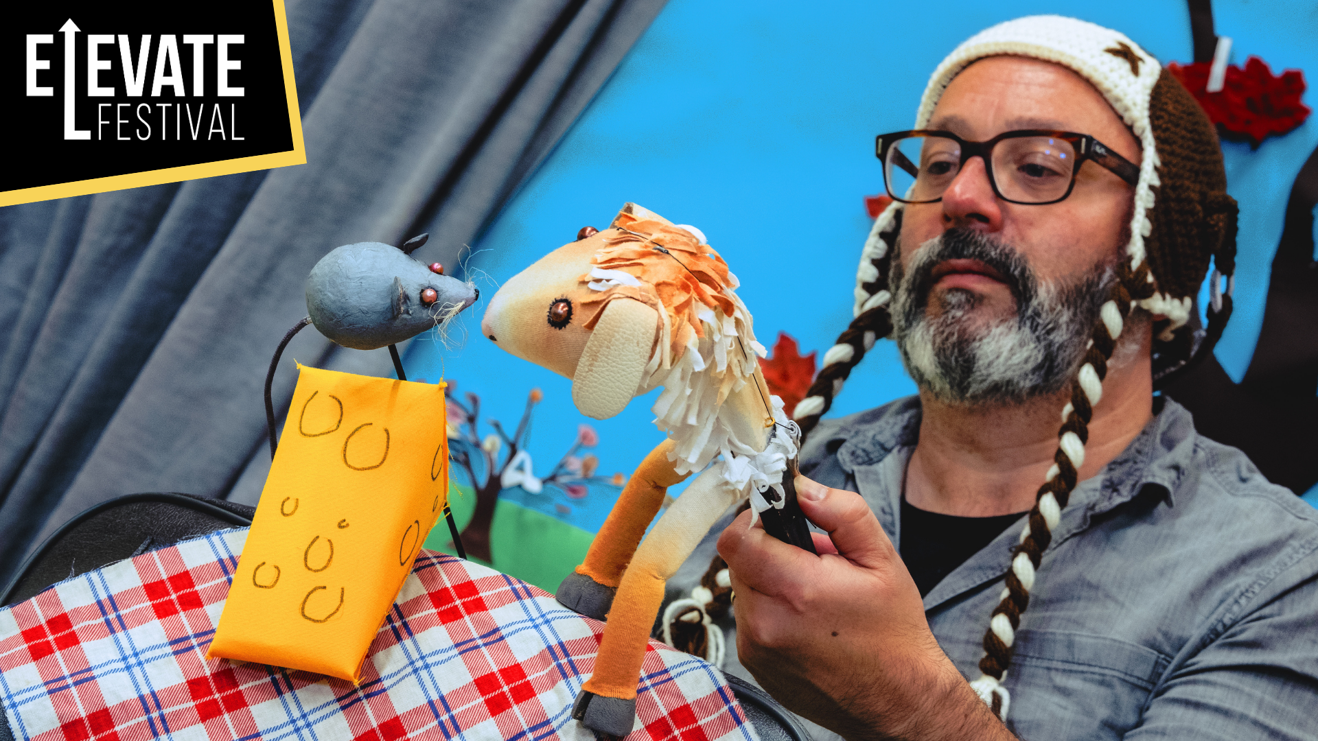 The Thing About Goats artwork. Shows a man with a knitted hat and beard, Holding a horse puppet. He is bringing the puppet close to a puppet of a mouse sitting on a triangle of cheese.