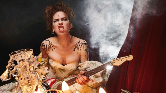 Credit: Matt Crockett. Emma Frankland is photographed in a dramatic theatrical setting, gripping a red electric guitar, directing an intense expression at camera. Wearing a voluminous off-the-shoulder floral gown, marked with bold black body paint across shoulders and chest. Warm chandelier and candle light in the foreground; theatrical smoke, a diagonal light beam and deep red stage curtains behind. Visible guitar cables and stage equipment.