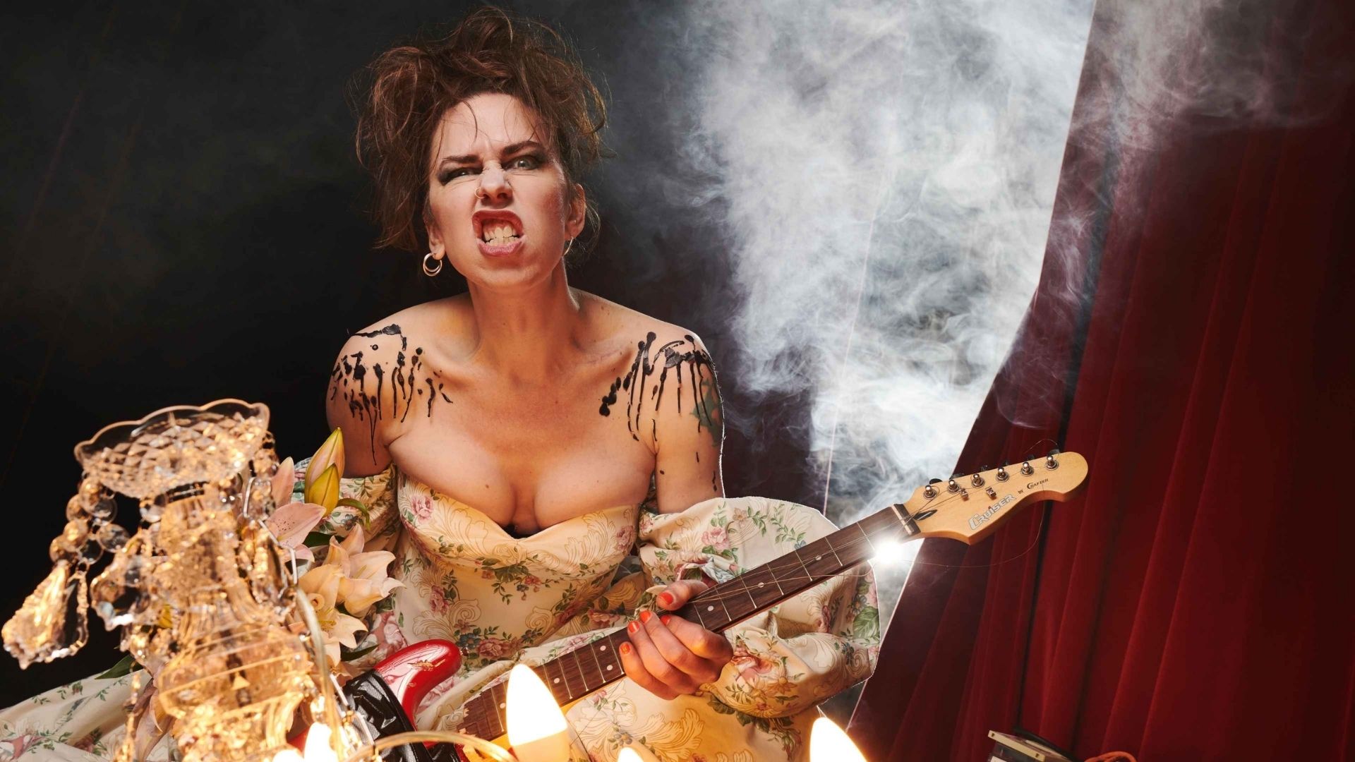 Credit: Matt Crockett. Emma Frankland is photographed in a dramatic theatrical setting, gripping a red electric guitar, directing an intense expression at camera. Wearing a voluminous off-the-shoulder floral gown, marked with bold black body paint across shoulders and chest. Warm chandelier and candle light in the foreground; theatrical smoke, a diagonal light beam and deep red stage curtains behind. Visible guitar cables and stage equipment.